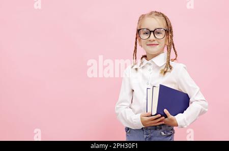 Ein bezauberndes Schulmädchen mit lustigen Zöpfen auf dem Kopf, das Lehrbücher in den Händen hält, in der Kamera lächelt und die Brille mit dem Finger auf pinkfarbenen Hintergrund fixiert, bereitet sich darauf vor Stockfoto