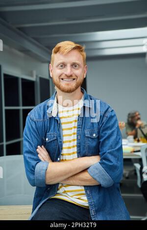 Vertikaler junger rothaariger Programmierer oder Geschäftsmann in einem modernen Büro, der wie eine Kamera aussieht und lächelt. Stockfoto