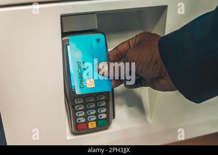 Kontaktloses Zahlungssystem. Ein Afro-Typ im Anzug, der eine Kreditkarte in der Nähe eines elektronischen Zahlungsautomaten hält, bezahlt für die Lieferung. Mann, der mit NFC technol bezahlt Stockfoto