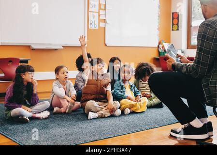 Der Junge hebt die Hand, um eine Frage in einem Klassenzimmer zu beantworten; er sitzt mit anderen Kindern auf dem Boden und der Lehrer sitzt vor der Klasse. Stockfoto