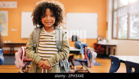 Junger männlicher Schüler, der lächelt und vor die Kamera schaut, am ersten Tag des Zweitunterrichts. Ein Grundschüler steht mit seiner Schultasche auf dem Rücken, bereit Stockfoto