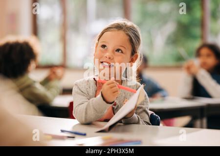 Aufgeregtes kleines Mädchen lernt in einem Kunstkurs mit einem Farbstift zu zeichnen. Fröhliche Grundschülerin spricht mit ihrem Lehrer, während sie einen Farbstift A hält Stockfoto