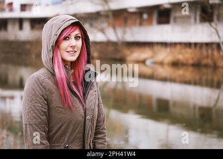 Durchbohrte junge Frau mit rosa Haaren und Kapuzenparka, die am Fluss steht Stockfoto