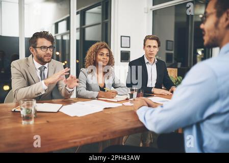 Sagen Sie uns, warum wir Sie einstellen sollten. Eine Gruppe von Geschäftsleuten, die einen Kandidaten in einem Büro interviewen. Stockfoto