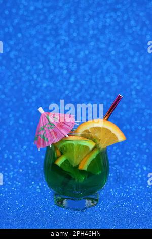 Ein kaltes, tropisches, grün gefärbtes Getränk in einem Glas mit orangefarbenen Scheiben, Limette und Sonnenschirm auf leuchtendem, leuchtend blauem Hintergrund Stockfoto