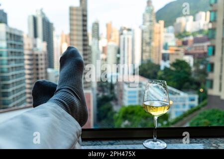 Mann entspannt und liegt auf einem Sessel mit Füßen auf der Fensterbank und Glas Wein. Verschwommene Stadtgebäude im Hintergrund Stockfoto