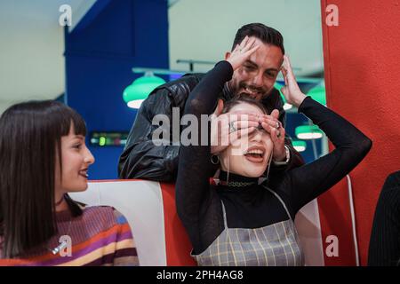 Ein weißer Mann verdeckt die Augen einer asiatischen Frau, während sie versucht zu erraten, wer er ist. Sie sind in einer Arkadenbar, mit einem lächelnden Teenager Freund, der im Sitzen sitzt Stockfoto