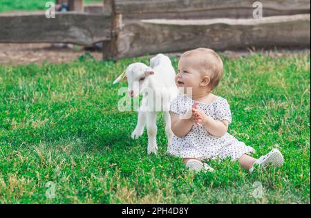 Ein wunderschönes Baby im Erbsenkleid spielt mit einer Ziege auf einem Bauernhof Stockfoto