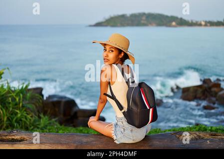 Eine multiethnische Frau mit Strohhut genießt einen tropischen Urlaub auf einer Bank an der Klippe mit Blick auf das Meer. Schwarze Frau mit Rucksack für Besichtigungstouren auf landschaftlich reizvoller Landschaft Stockfoto