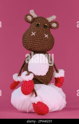 Amigurumi ein brauner Stier trägt rote Fäustlinge und Hausschuhe und sitzt auf einem weißen Fadenball auf pinkfarbenem Hintergrund. Weiches Heimwerkerspielzeug aus natürlicher Baumwolle und Stockfoto