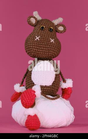 Amigurumi ein brauner Stier trägt rote Fäustlinge und Hausschuhe und sitzt auf einem weißen Fadenball auf pinkfarbenem Hintergrund. Weiches Heimwerkerspielzeug aus natürlicher Baumwolle und Stockfoto