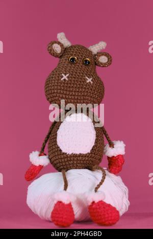 Amigurumi ein brauner Stier trägt rote Fäustlinge und Hausschuhe und sitzt auf einem weißen Fadenball auf pinkfarbenem Hintergrund. Weiches Heimwerkerspielzeug aus natürlicher Baumwolle und Stockfoto