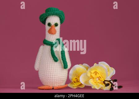 Amigurumi-Entenpuppe auf pinkfarbenem Hintergrund mit gelben Orchideenblumen. Weiches Heimwerkerspielzeug aus Baumwolle. Eine weiße Gans mit orangefarbenem Schnabel trägt einen grünen Hut und Stockfoto
