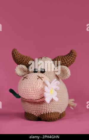 Amigurumi-Kuhpuppe auf pinkfarbenem Hintergrund. Ein weiches Heimwerkerspielzeug aus Baumwolle und Naturwolle. Ein brauner Stier mit einer Gänseblümchen im Mund, gehäkert, handgemacht Stockfoto