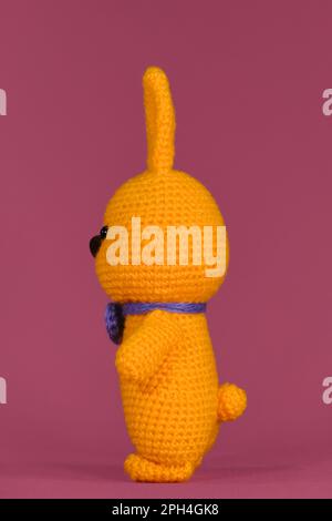 Orangefarbenes Osterhasen mit lila Fliege, gestrickt, handgefertigt. Amigurumi Hase Puppe auf pinkfarbenem Hintergrund. Weiches Spielzeug aus Baumwolle und Wolle. Symbo Stockfoto