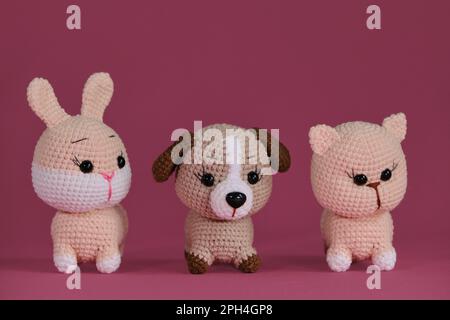 Amigurumi drei Tierpuppen auf pinkfarbenem Hintergrund. Heimwerker-Spielzeug aus natürlicher Baumwolle und Wolle. Vorderansicht. Süßes kleines Kätzchen, Hündchen und Häkelchen Stockfoto