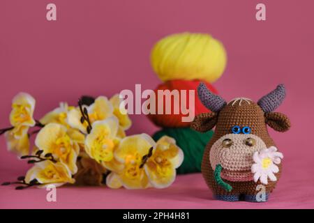 Ein brauner Stier mit Gänseblümchen im Mund, gestrickt, handgemachte Kunst. Amigurumi-Kuhpuppe auf pinkfarbenem Hintergrund neben gelben Orchideenblumen und mehrfarbiger Wolle Stockfoto