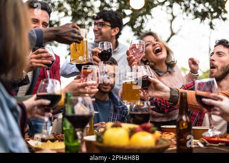 Eine vielfältige Gruppe von Freunden, die bei einem Picknick auf dem Land ihre Gläser erheben. Der Tisch ist mit gegrilltem Fleisch, Wein und Bier umgeben Stockfoto