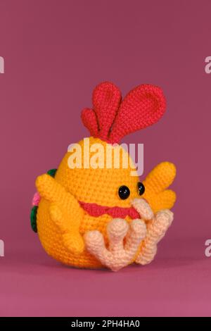 Orangenhahn mit leuchtend rotem Kamm auf dem Kopf, gehäckselte, handgemachte Kunst. Amigurumi Puppe Huhn mit bunten Schwänzen auf pinkfarbenem Hintergrund. Weiches Heimwerkerprodukt Stockfoto