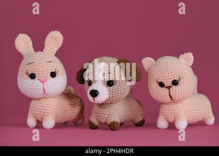 Amigurumi drei Tierpuppen auf pinkfarbenem Hintergrund. Heimwerker-Spielzeug aus natürlicher Baumwolle und Wolle. Vorderansicht. Süßes kleines Kätzchen, Hündchen und Häkelchen Stockfoto