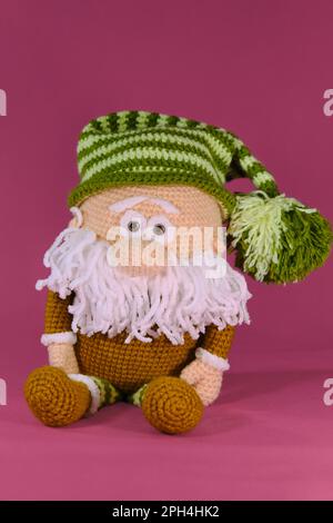 Amigurumi ein Zwerg mit grün gestreiftem Hut und weißem Bart auf pinkfarbenem Hintergrund. Gestrickter alter Mann, handgemachte Kunst. Weiches Heimwerkerspielzeug aus natürlicher Baumwolle Stockfoto
