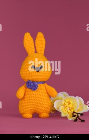 Symbol für katholische Ostern. Orangefarbenes Osterhasen mit lila Fliege, gestrickt, handgefertigt. Amigurumi Hase Puppe auf pinkfarbenem Hintergrund mit Gelb Stockfoto