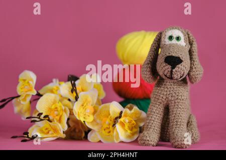 Amigurumi Puppe auf pinkfarbenem Hintergrund neben gelben Orchideenblumen und bunten Wollkugeln. Ein weiches Heimwerkerspielzeug aus Baumwolle. Ein brauner Hund Stockfoto