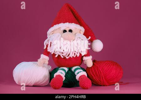 Amigurumi ein Weihnachtszwerg mit rotem Hut und weißem Bart auf pinkfarbenem Hintergrund. Weiches Spielzeug aus natürlicher Baumwolle und Wolle. Gestrickter weihnachtsmann Stockfoto