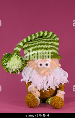 Amigurumi ein Zwerg mit grün gestreiftem Hut und weißem Bart auf pinkfarbenem Hintergrund. Gestrickter alter Mann, handgemachte Kunst. Weiches Heimwerkerspielzeug aus natürlicher Baumwolle Stockfoto