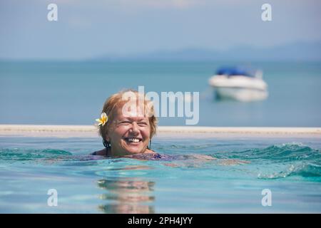 Aktive, lachende Seniorin im Pool gegen das Meer. Die Frau schwimmt an sonnigen Tagen. Stockfoto