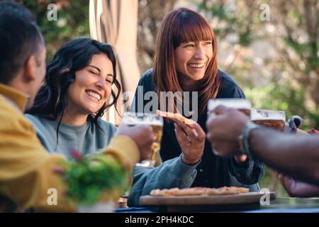 Eine Gruppe junger Millennials-Freunde, die sich an einem Tisch im Freien amüsieren - Pizza essen und Bier trinken, während sie lachen und reden - brünett Stockfoto