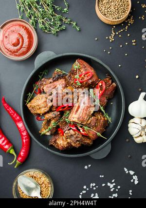 Gebackene Rippchen in einer Pfanne. Gegrillte Schweinerippchen mit Gewürzen und Kräutern auf dunklem Hintergrund. Lebensmittelhintergrund. Draufsicht. Stockfoto