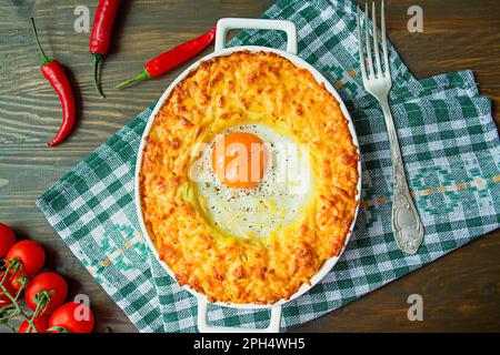 Kartoffelauflauf mit Bolognese. Auflauf für gebackene Kartoffeln mit Ei und geriebenem Käse in einem keramischen, ovalen Backblech. Dunkler Holzhintergrund. Stockfoto