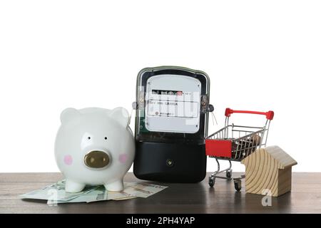 Stromzähler, Hausmodell, Sparschwein und Einkaufswagen mit Geld auf Holztisch vor weißem Hintergrund Stockfoto