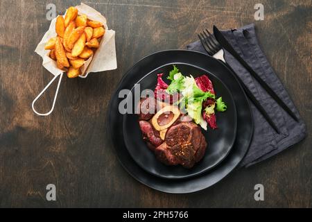 Steaks, Osso Buco. Rinderscheiben, gegrillter Osso Buco oder Porterhouse-Fleischsteak mit garniertem Salat und pommes frites auf schwarzem Marmorbrett auf altem Holz Stockfoto