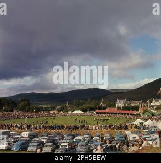 1960er, historisch, Blick auf die Highland Games in Braemar, Schottland, Großbritannien, mit Autos und Zuschauern und den Bergen im Hintergrund. Auch bekannt als Braemer Gathering, sind die Spiele die meistbesuchten Highland-Spiele in Schottland. Stockfoto