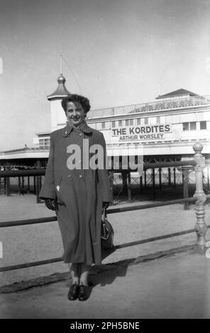 1954, historisch, eine Städterin in einem langen Mantel, die vor dem Central Pier in der Küstenstadt Blackpool, Lancashire, England, für ein Foto steht. In diesem Jahr erschienen dort die Korditen und Arthur Worsley, deren Namen an der Seite des Pier-Gebäudes stehen. Arthur Worsley war ein beliebter britischer Bauchredner der Epoche mit regelmäßigen Fernsehauftritten. Die Korditen waren eine männliche und weibliche Gesangsgruppe, die auch Skizzen mit Liedern aufführte und eine regelmäßige Nummer im BBC-Lichtprogramm „Variety Fanfare“ war. Stockfoto