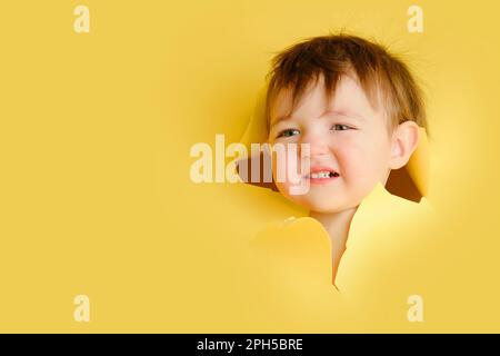 Wütendes Baby in einem Loch auf papiergelbem Hintergrund. Zerrissener Studiohintergrund des Kindes, Kopierraum. Ein Kind im Alter von einem Jahr, sechs Monaten Stockfoto