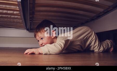 Das Baby krabbelt und versteckt sich unter dem Bett. Das Kind kletterte unter das Sofa und setzte sich auf den Boden. Ein Junge im Alter von einem Jahr, acht Monate, volle Länge Stockfoto