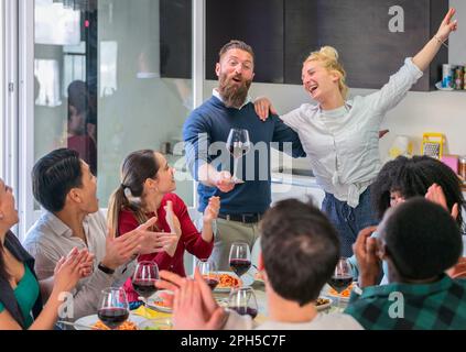 Junge, multiethnische Freunde sitzen am Tisch, feiern Thanksgiving, essen zu Hause zusammen und toasten, trinken, Gastrede halten mit Wein Stockfoto