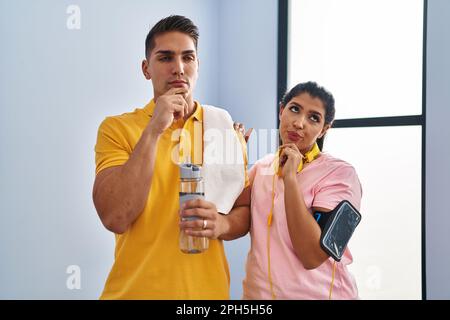 Ein junges Paar, das Sportbekleidung und Kopfhörer trägt, denkt ernsthaft mit der Hand am Kinn über Fragen nach, denkt über verwirrende Ideen nach Stockfoto