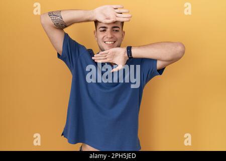 Junger hispanischer Mann, der auf gelbem Hintergrund lächelt und fröhlich einen Buh mit Händen und Gesicht zeigt, ist überrascht und begeistert Stockfoto