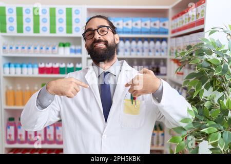 Ein hispanischer Mann mit Bart, der in der Apotheke arbeitet, der selbstbewusst aussieht, mit einem Lächeln im Gesicht, mit stolzen Fingern und glücklich auf sich zeigt. Stockfoto