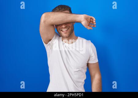 Junger weißer Mann, der auf blauem Hintergrund steht und die Augen mit einem lächelnden Arm bedeckt, fröhlich und lustig. Blindes Konzept. Stockfoto