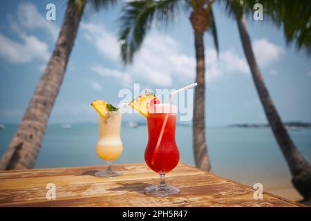 Zwei Cocktails unter Palmen am wunderschönen Strand. Singapore Sling und Pina Colada Drinks auf Holztisch. Stockfoto