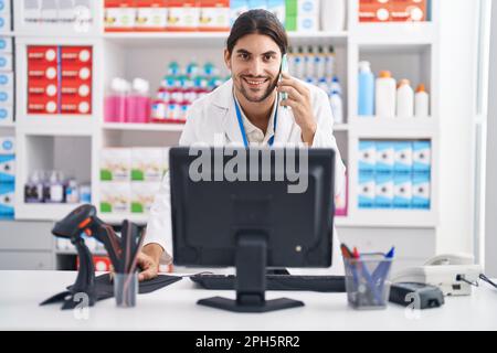 Junger hispanic-Mann Apotheker im Gespräch auf Smartphone mit Computer in der Apotheke Stockfoto