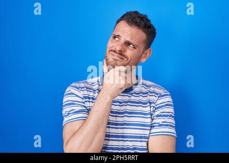Junger hispanischer Mann, der auf blauem Hintergrund steht und sich Sorgen um eine Frage macht, besorgt und nervös mit der Hand am Kinn Stockfoto