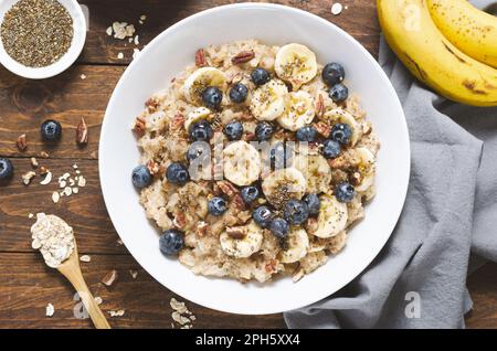 Haferflocken-Schale, Haferbrei mit Blaubeeren, Bananen und Pekannüsse in einer Schüssel auf rustikalem Holzhintergrund, gesunder Snack oder Frühstück Stockfoto