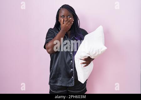 Junge afrikanische Frau, die Pijama trägt, Kissen umarmt, den Mund bedeckt, mit Händen für einen Fehler. Geheimes Konzept. Stockfoto