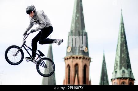 Oldenburg, Deutschland. 26. März 2023. Luca Siedek springt während einer Vorführung über eine Rampe auf seinem BMX-Fahrrad, während die Türme der Lamberti-Kirche im Stadtzentrum im Hintergrund zu sehen sind. Mit verschiedenen Aktionen wie einer BMX-Show, einer Fahrradcodierung, einer Reinigungsaktion für Fahrräder oder der Auktion von Zweirädern klingelt die Fahrradstadt Oldenburg im Frühjahr. Kredit: Hauke-Christian Dittrich/dpa/Alamy Live News Stockfoto